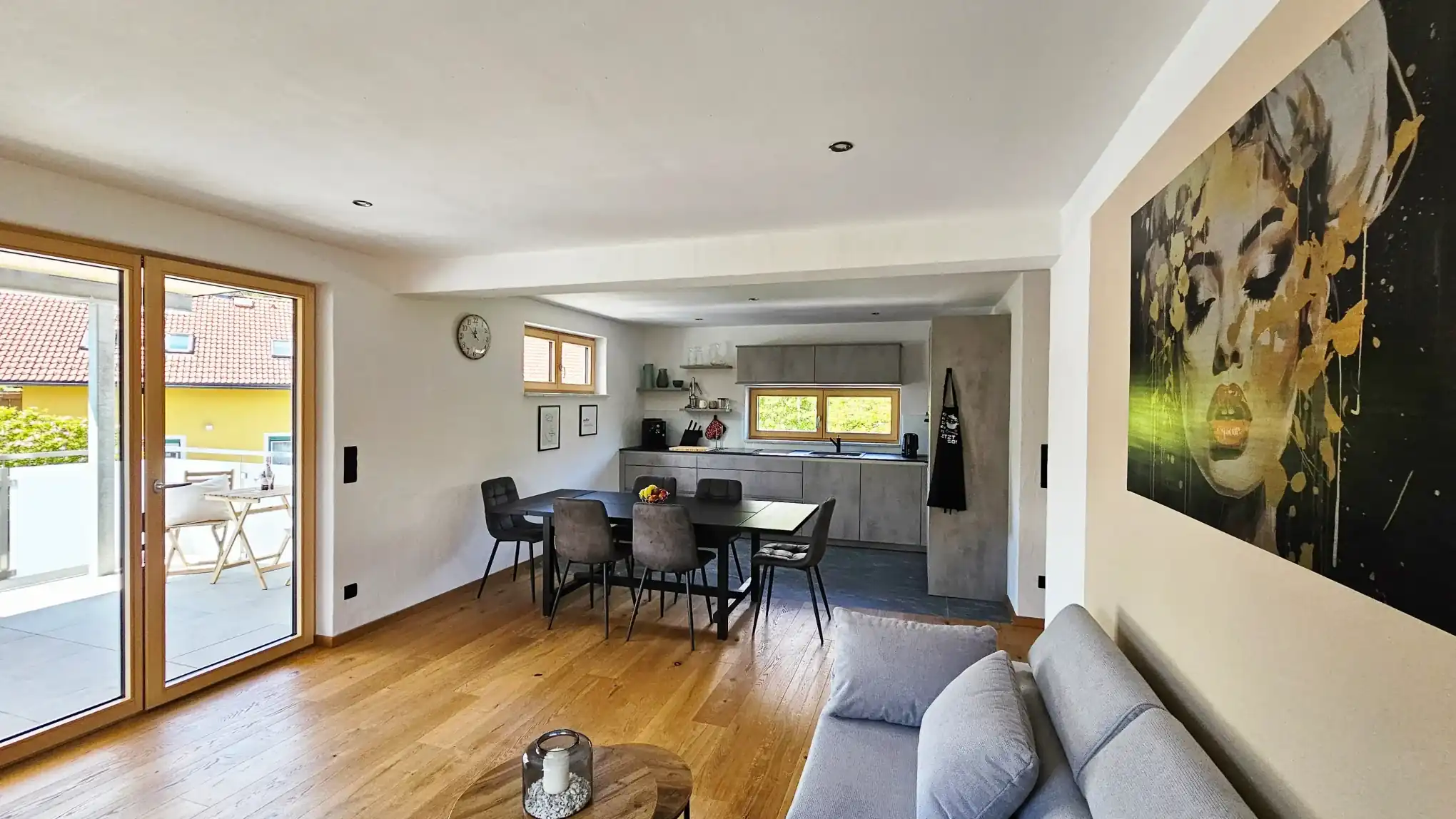 Heller offener Wohn- und Essbereich mit Holzböden, einem grauen Sofa, einem schwarzen Esstisch mit vier Stühlen und einer modernen grauen Küche im Hintergrund. Große Glastüren öffnen sich zu einem Balkon, und ein großes Porträt hängt an der rechten Wand.