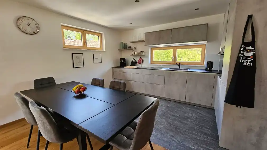 Offene Wohnküche mit einem großen schwarzen Esstisch, sechs gepolsterten grauen Stühlen und einer Obstschale in der Mitte. Im Hintergrund eine moderne graue Küchenzeile mit dunkler Arbeitsplatte, Spüle, Kaffeemaschine und Messerblock. Zwei Holzrahmenfenster lassen natürliches Licht herein, an der linken Wand hängt eine Vintage-Wanduhr, rechts eine schwarze Kochschürze.