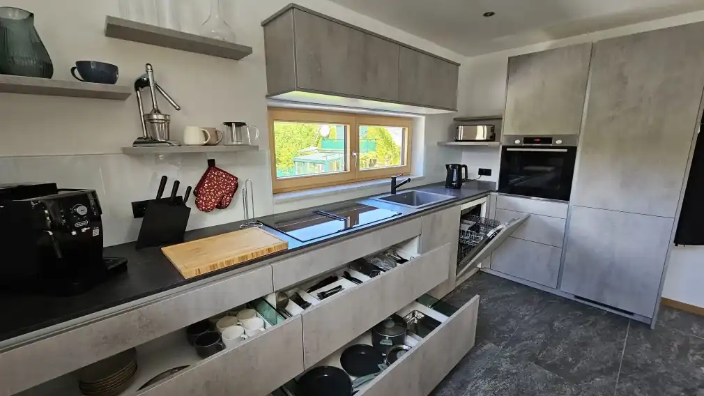 Moderne Küche mit grauen Schränken, offenen Schubladen, die organisierte Geschirr- und Kochutensilien zeigen, einem eingebauten Ofen, einer geöffneten Spülmaschine und einer Arbeitsfläche mit Kaffeemaschine, Schneidebrett und Utensilien. Ein Fenster sorgt für natürliches Licht über der Spüle.