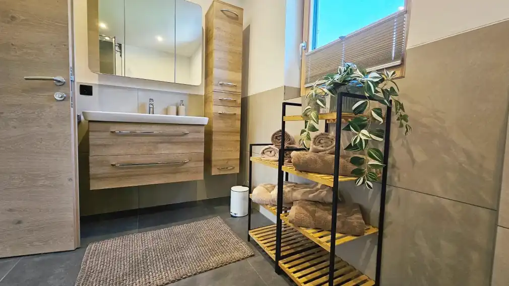 Modernes Badezimmer mit Holzschränken, einem weißen Waschbecken und einem spiegelnden Medizinschrank. Rechts befindet sich ein schwarzes Metall- und Holzregal, das ordentlich aufgerollte braune Handtücher und eine grüne Topfpflanze hält. Ein gewebter Teppich liegt auf dem dunklen Fliesenboden, und Licht strömt durch ein Fenster mit Plisseerollo.
