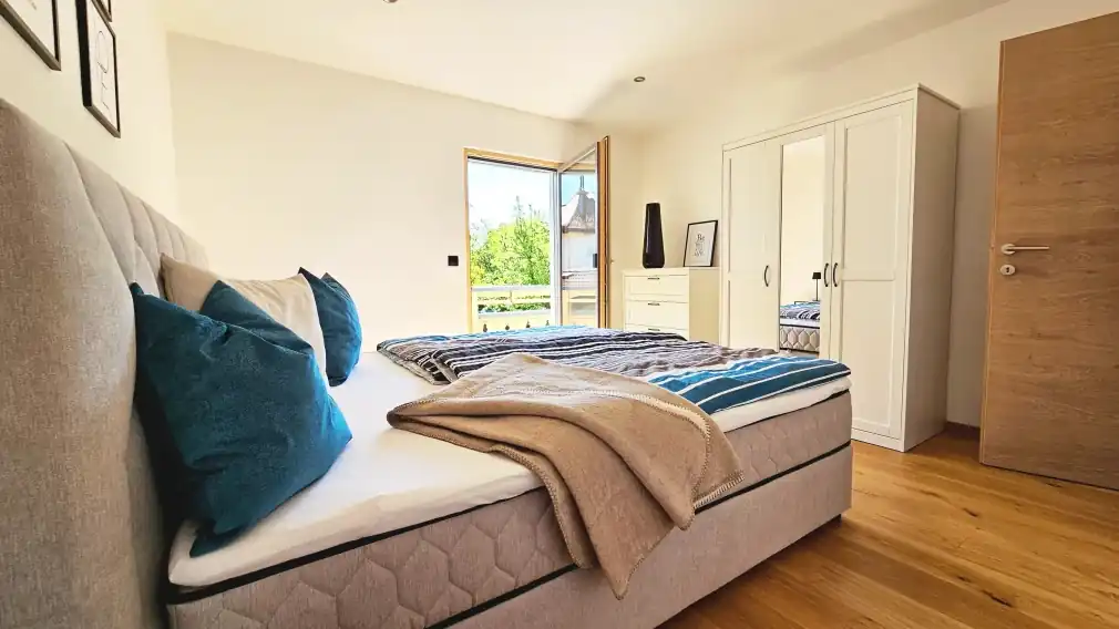 Detailaufnahme des sonnigen Schlafzimmers mit einem beigefarbenen, gepolsterten Boxspringbett, türkis- und beigefarbener gestreifter Bettwäsche und mehreren Zierkissen, gerahmten Bildern an der Wand und einer gläsernen Balkontür.