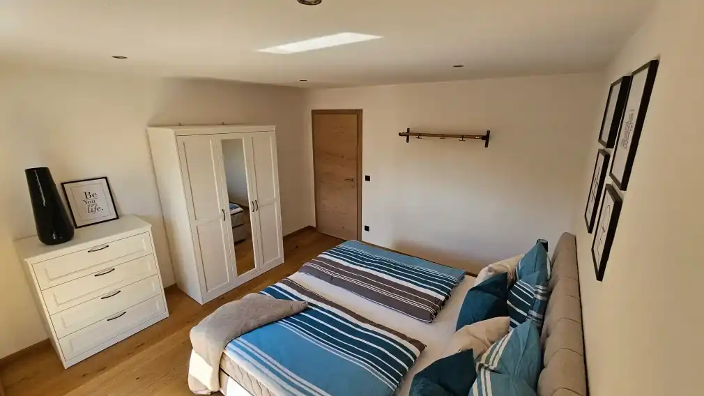 Detailaufnahme des sonnigen Schlafzimmers mit einem beigefarbenen, gepolsterten Boxspringbett, türkis- und beigefarbener gestreifter Bettwäsche und mehreren Zierkissen gerahmten Bildern an der Wand.