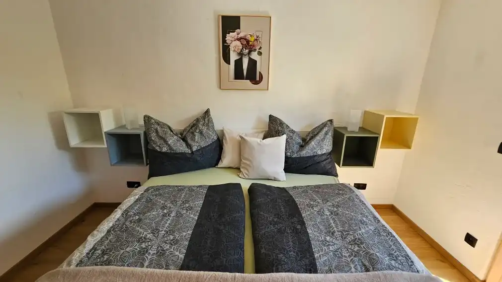 Detailaufnahme des kleinen, hellen Schlafzimmers mit einem weißen Plattformbett, grauen, gemusterten Bettdecken und pastellfarbenen Kissen, geometrischen Wandregalen und einem gerahmten Blumenbild über dem Bett.