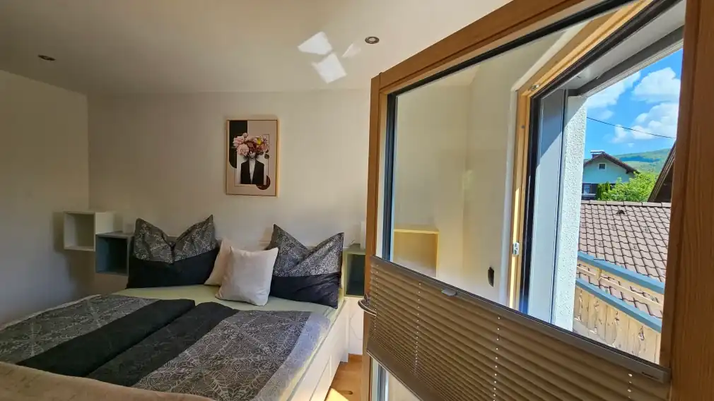 Detailaufnahme des kleinen, hellen Schlafzimmers mit einem weißen Plattformbett, grauen, gemusterten Bettdecken und pastellfarbenen Kissen, geometrischen Wandregalen und einem gerahmten Blumenbild über dem Bett.