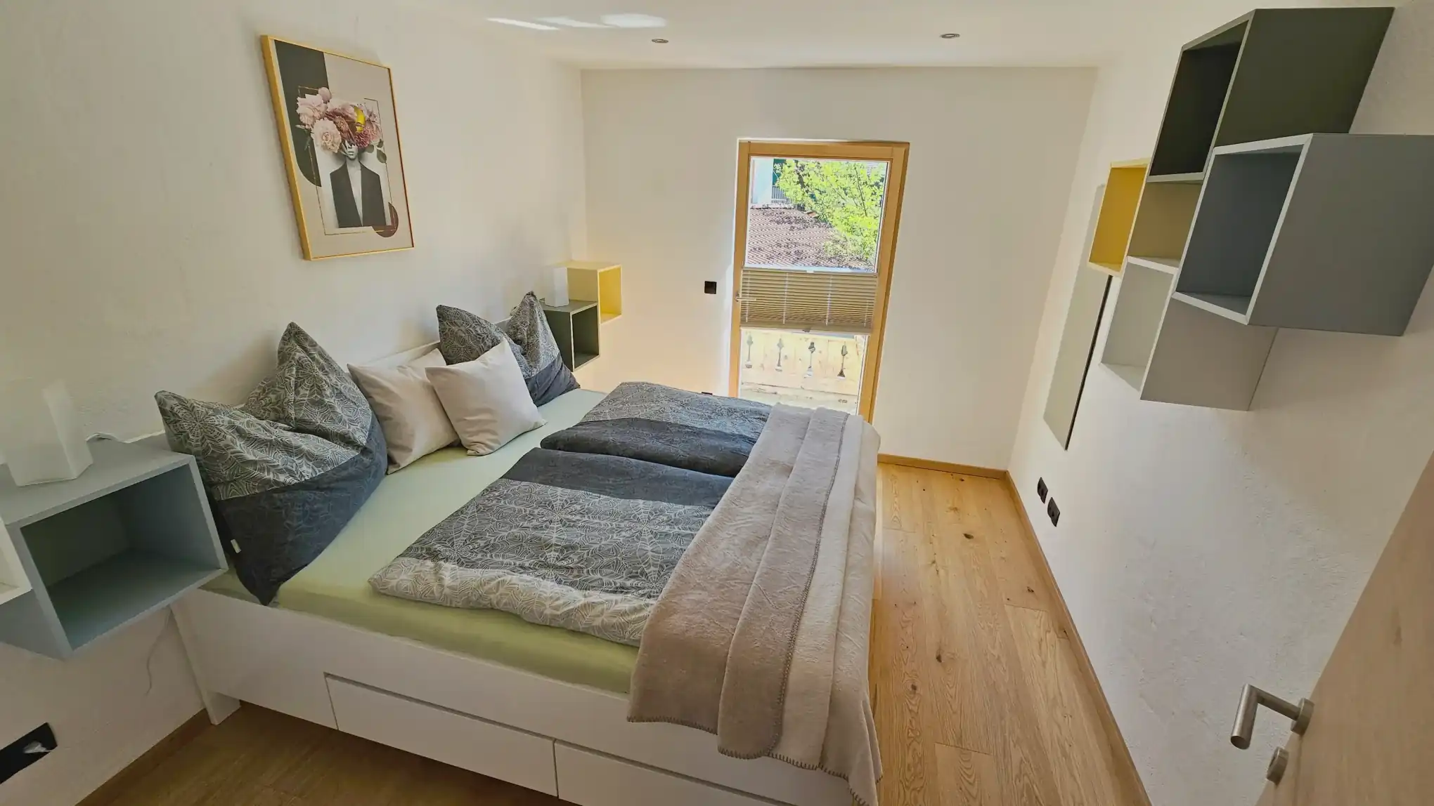 Kleines, helles Schlafzimmer mit einem weißen Plattformbett, das mit grauen, gemusterten Bettdecken und pastellfarbenen Kissen ausgestattet ist, geometrischen Wandregalen, einem gerahmten Blumenbild über dem Bett, einer Glastür zu einem kleinen Balkon und hellen Eichenholzböden.