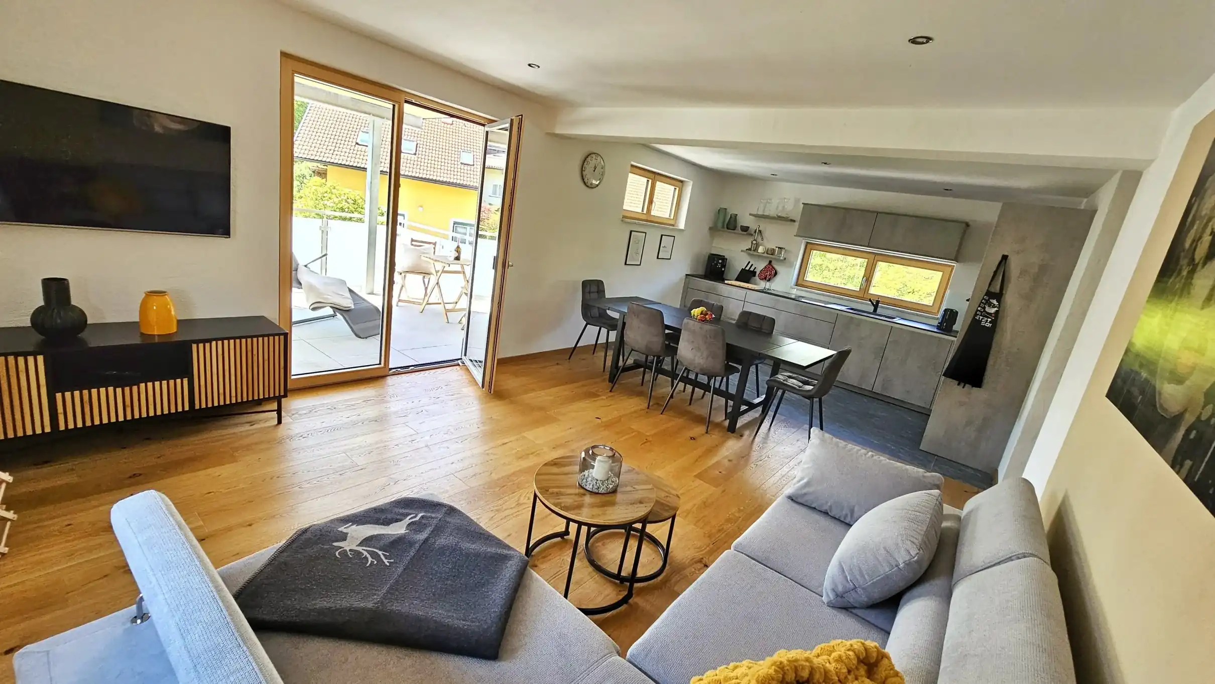Heller, offener Wohn- und Essbereich mit Eichenholzböden, einem grauen Ecksofa mit Zierkissen und Hirschdecke, einem runden Couchtisch, einem TV-Sideboard aus Holz und Metall sowie einem Flachbildfernseher an der Wand. Im Hintergrund ein schwarzer Esstisch mit gepolsterten Stühlen und eine moderne graue Küchenzeile. Eine geöffnete Glastür führt auf einen sonnigen Balkon mit Blick auf die Nachbarschaft. An der rechten Wand hängt ein großes Landschaftsbild.